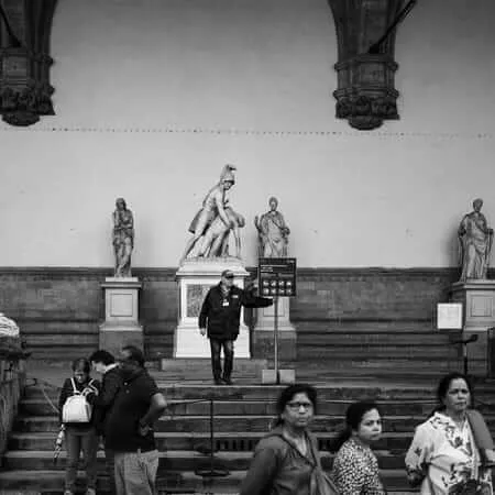 Black and white photograph of tourists gathered at the Loggia dei Lanzi in Florence, Italy, where a security guard stands on the steps pointing toward an information sign, surrounded by Renaissance sculptures including lions and classical figures, while visitors of diverse backgrounds mill about in the foreground.