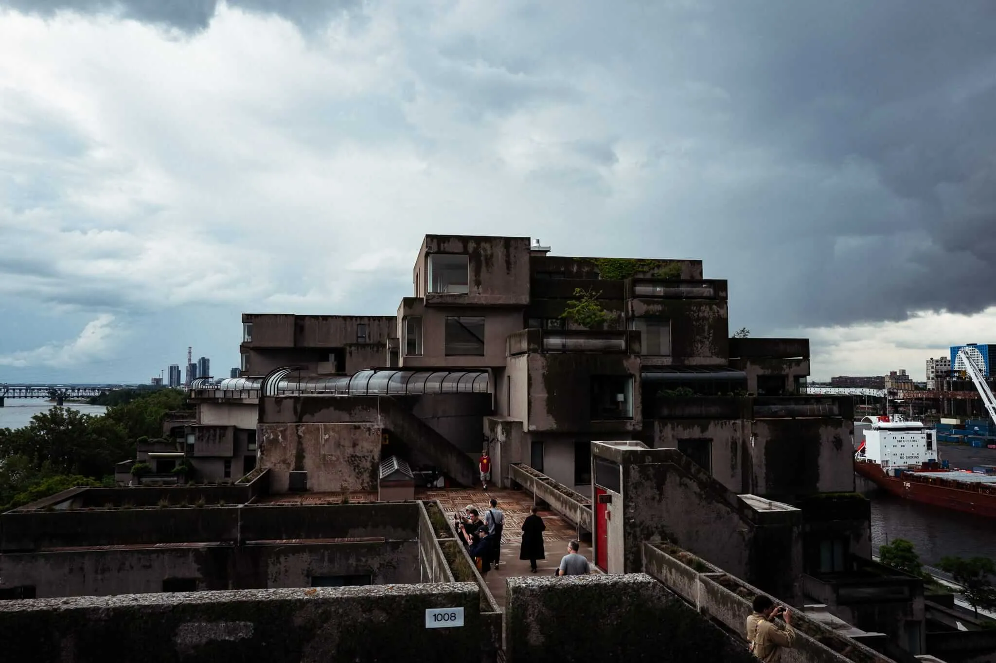 Visitors explore the weathered concrete terraces and walkways of Habitat 67 in Montreal, with a dramatic stormy sky overhead and the St. Lawrence River and port visible in the background.
