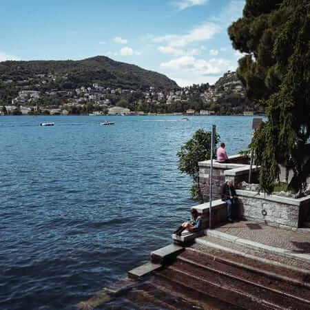 People resting on stone steps and walls along a lakeside promenade, with boats on the water and a hillside town visible across the lake under a partly cloudy sky