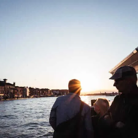 Passengers on a vaporetto water bus silhouetted against a blazing sunset over a Venetian canal, with historic buildings lining the waterway and warm golden light reflecting off the water