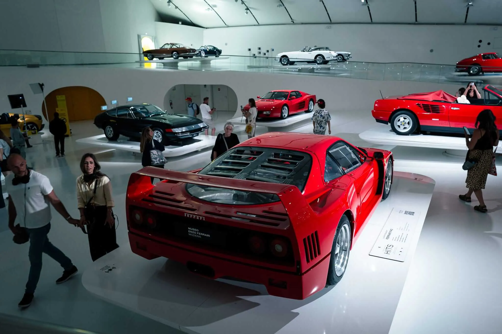 Interior of a modern Ferrari museum with multiple levels displaying classic and contemporary Ferrari sports cars. Visitors walk among red Ferrari models on white platforms in a minimalist white gallery space with dramatic lighting. The architecture features clean lines and geometric design, creating a temple-like atmosphere for automotive appreciation.