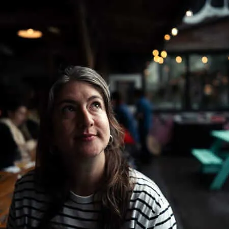 A woman with dark hair streaked with grey, wearing a white and black striped long-sleeve shirt, sits at a wooden table in a dimly lit outdoor venue. She gazes upward with a contemplative expression, illuminated by dramatic side lighting. Warm string lights and blurred figures populate the background.