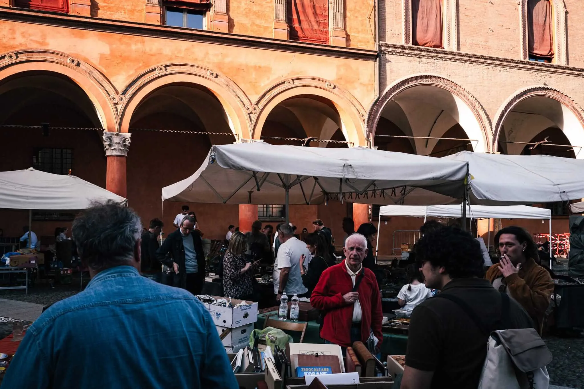 A bustling outdoor market scene under historic Renaissance arches with warm terracotta-colored walls. Multiple people browse market stalls with white canopies, including an elderly man in a red cardigan prominently featured in the center. The architecture features classical columns and arched openings typical of Italian piazzas.