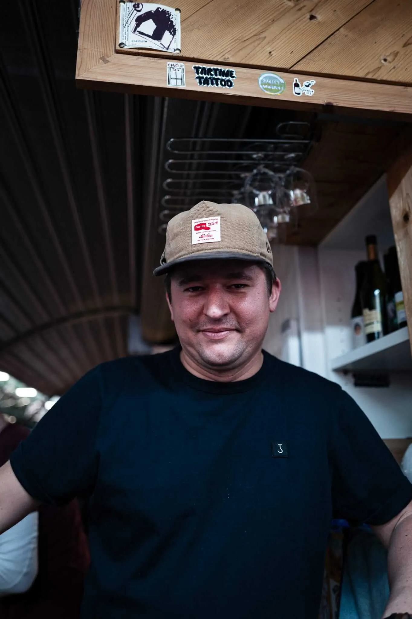 A middle-aged man in a tan baseball cap and dark t-shirt smiles warmly at the camera while standing in what appears to be a rustic restaurant or bar. Behind him, wooden beams display various stickers including 'Take Time Tattoo' and other local business logos. Wine glasses hang from a rack above, and the warm wood-paneled interior suggests a cozy, community-oriented establishment.