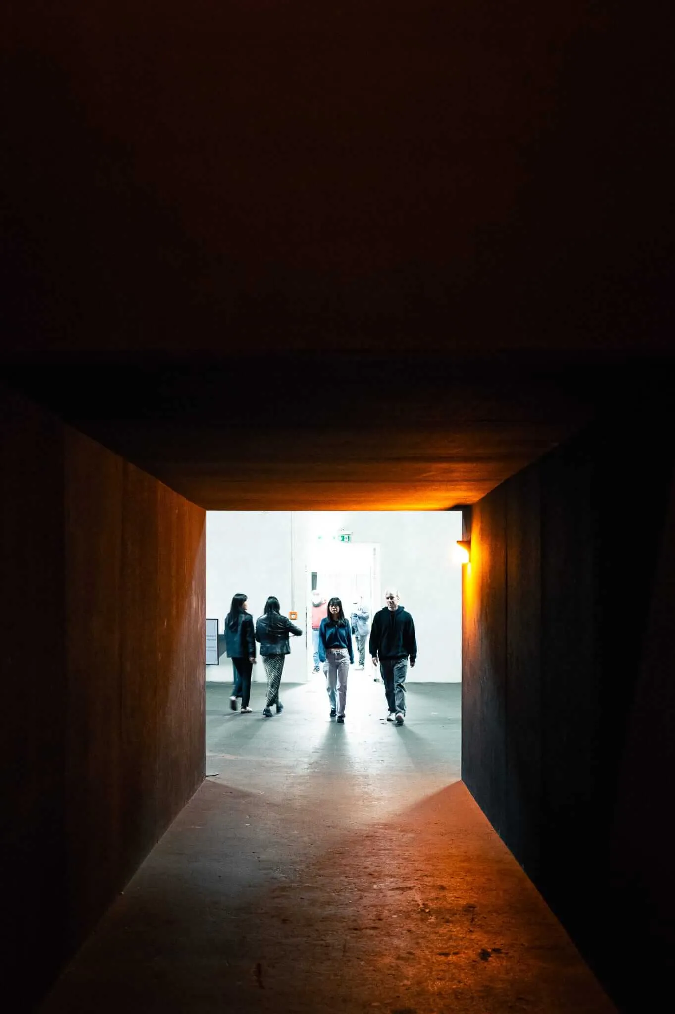 A group of people walking through a dark tunnel or underpass toward bright daylight at the far end. The tunnel walls are concrete, and warm orange light illuminates the ceiling and upper walls, creating dramatic contrast with the bright white light visible at the tunnel's exit where silhouetted figures can be seen moving.