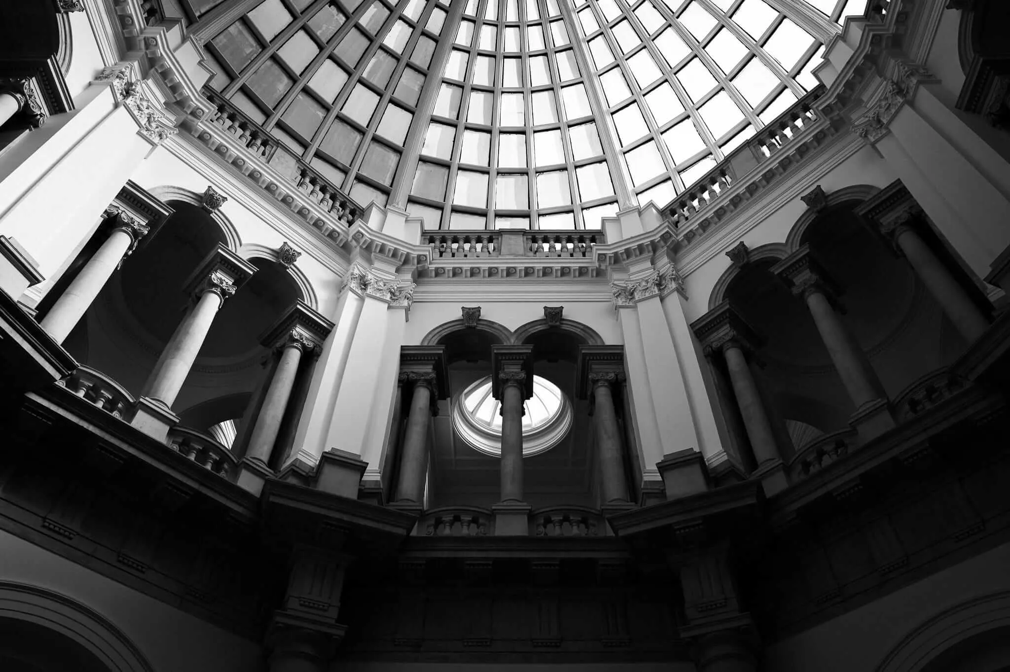 Black and white upward view of a classical domed interior featuring ornate Corinthian columns, decorative molding, and a dramatic circular glass ceiling with geometric grid pattern letting in bright natural light