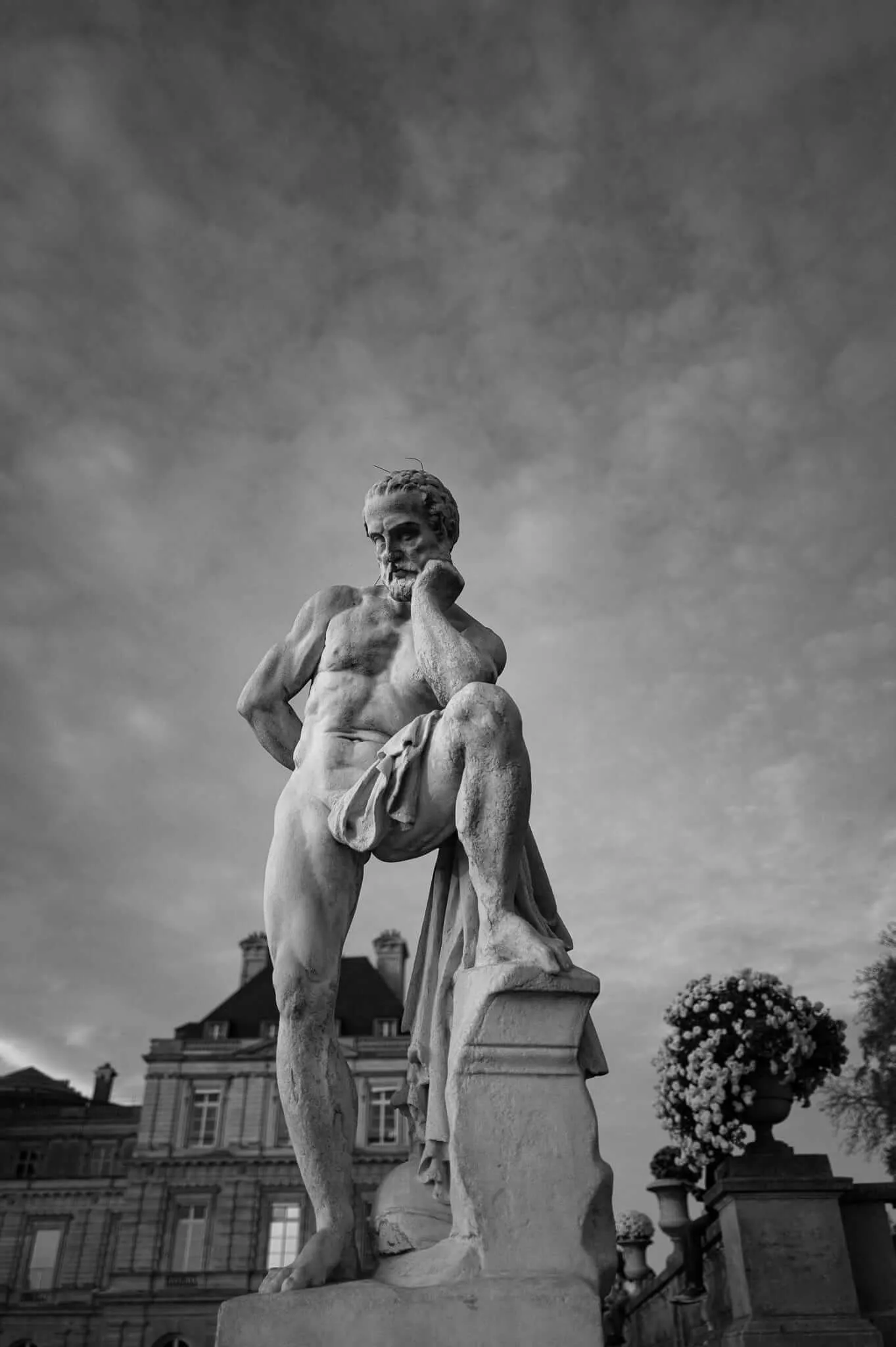 Black and white photograph of a classical marble statue depicting a bearded male figure in a contemplative pose with hand to chin, shot from below against a dramatic cloudy sky with historic architecture visible in the background