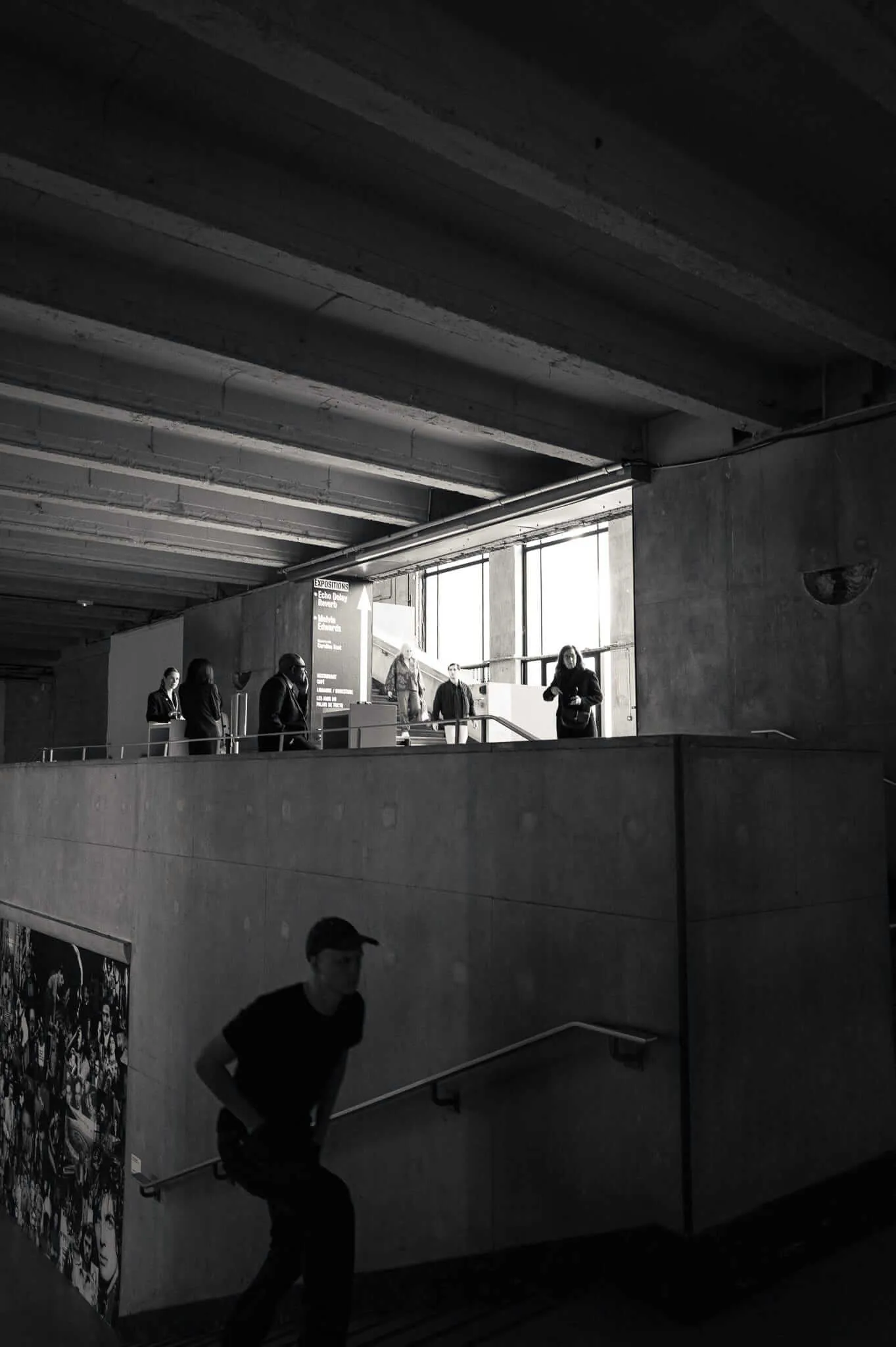 Black and white photograph showing a multi-level concrete interior space with exposed ceiling beams. Several people stand on an upper level near windows and directional signage, while a solitary figure in dark clothing sits on stairs in the lower level. The stark brutalist architecture creates dramatic shadows and geometric forms.