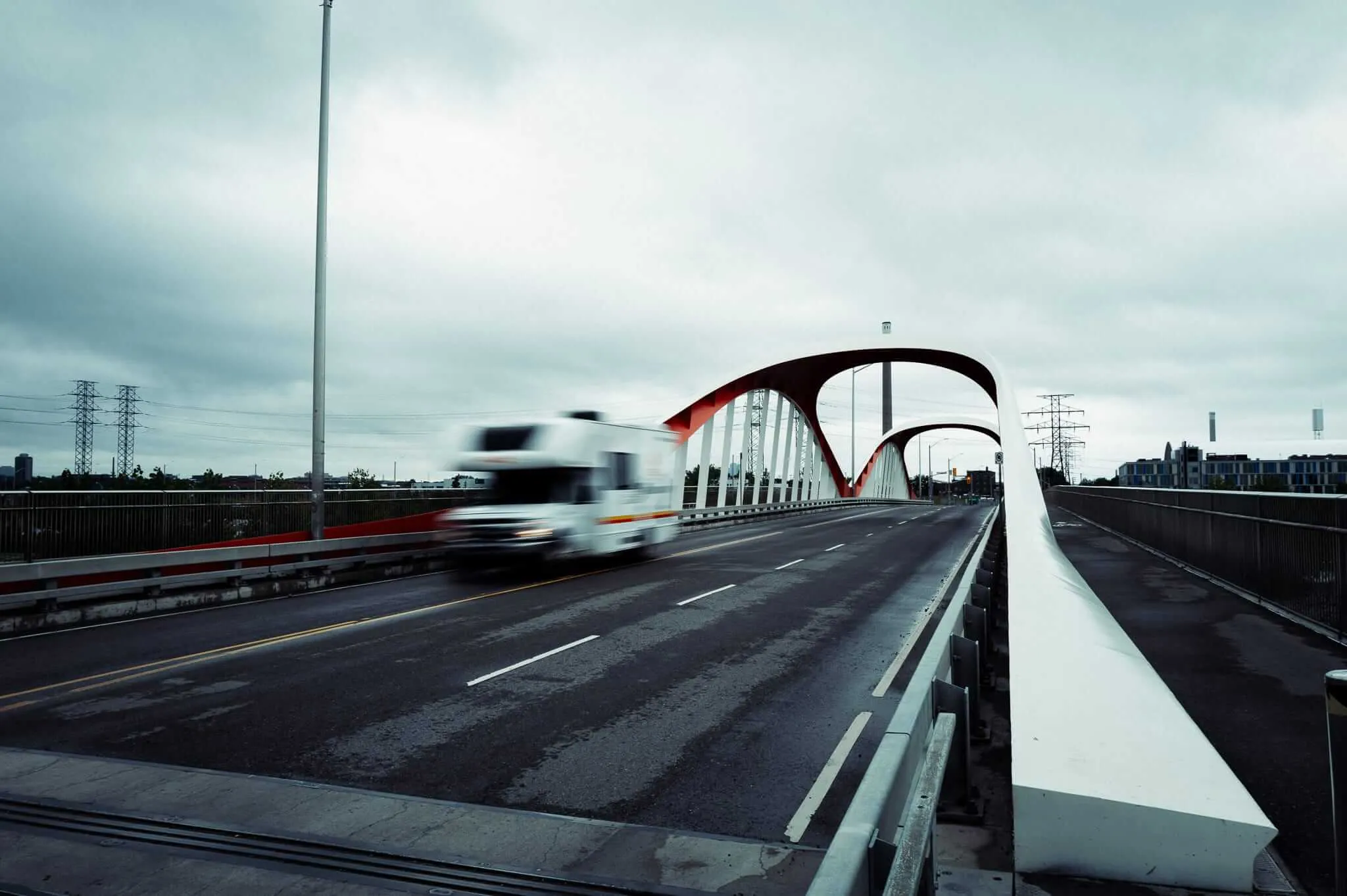 A motion-blurred white transport truck crosses a modern arch bridge with striking red and white steel arches under an overcast grey sky, with power transmission towers visible in the background.