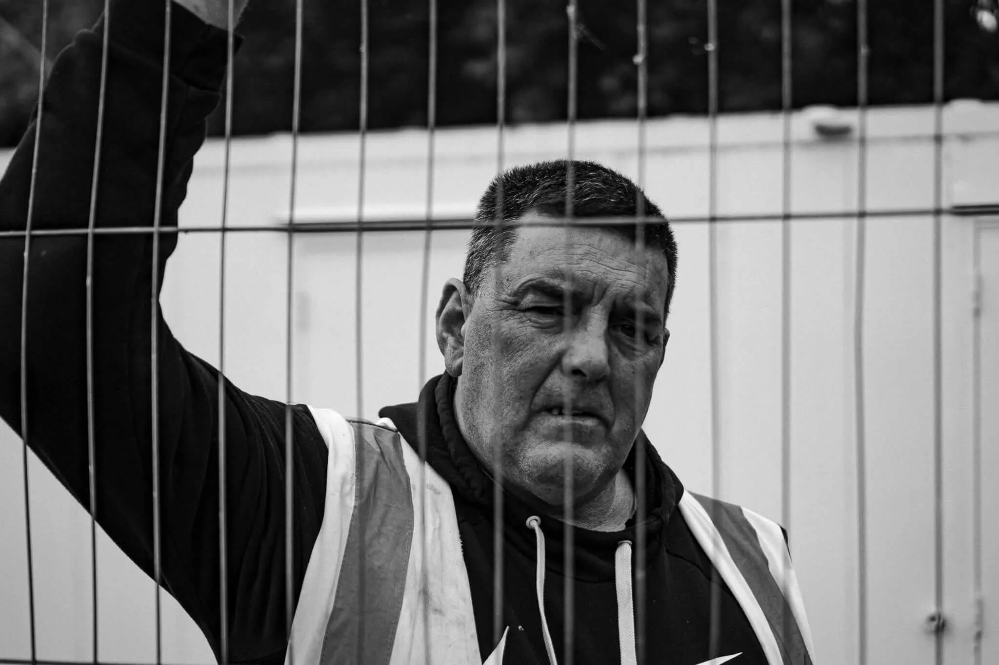 A middle-aged man in a high-visibility safety vest grips a wire mesh fence with one raised hand, gazing intensely into the distance. The black and white portrait is shot close-up through the fence, with a white structure visible in the background.