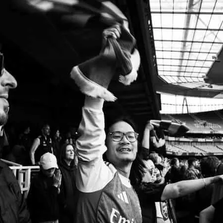 Black and white photograph of excited sports fans in a stadium, with a man in glasses and scarf celebrating with raised hands while another fan in a cap looks on, stadium architecture visible in the background