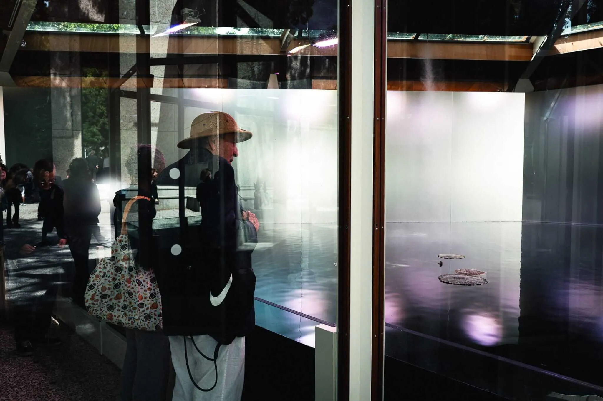 An elderly person wearing a straw hat and carrying a Nike bag and patterned tote bag stands before a glass exhibition case containing a large photographic artwork depicting lily pads on still water, their reflection merging with the artwork behind the glass.