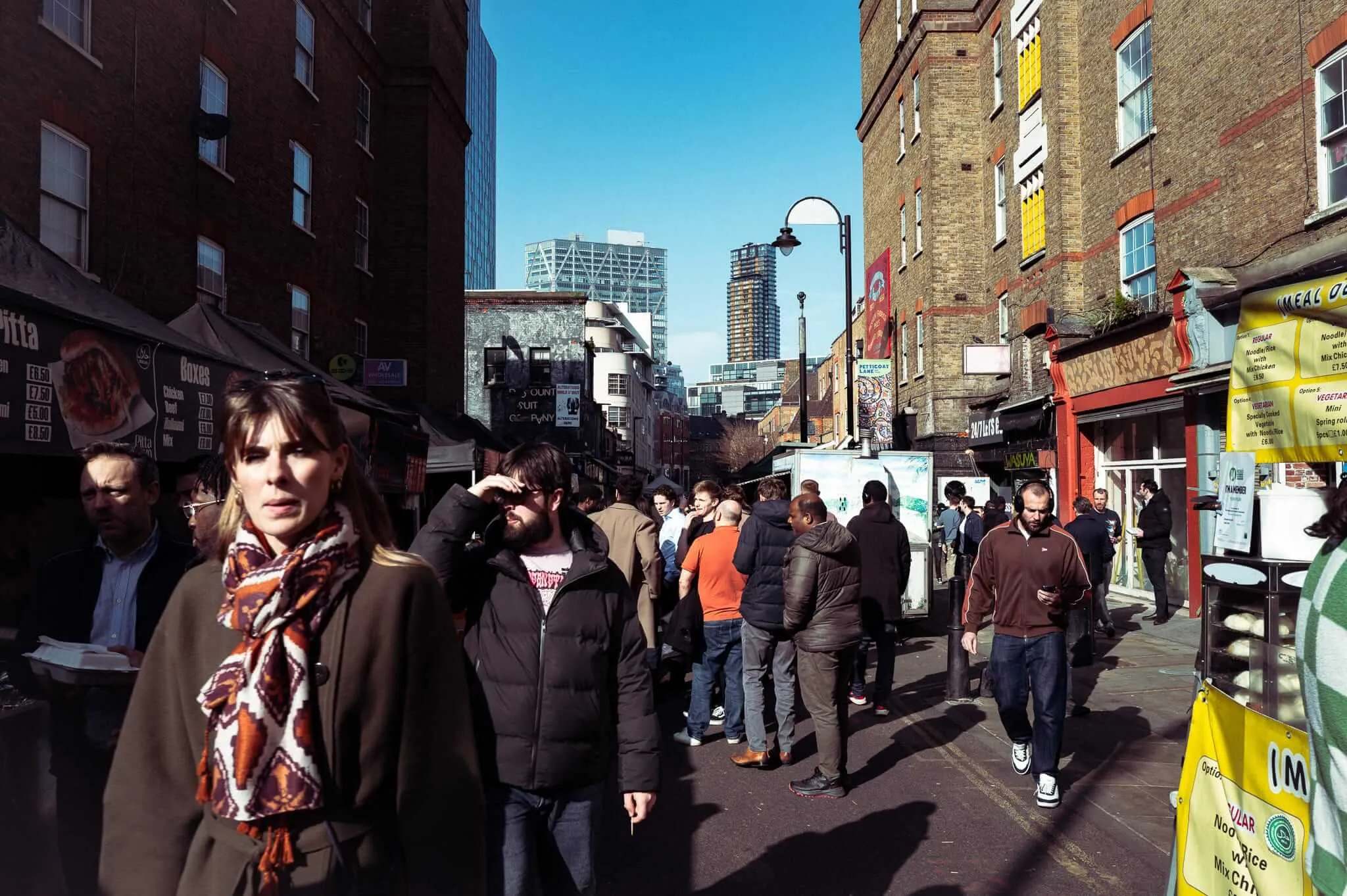 A bustling street market scene with crowds of people gathered around food stalls and vendors on a busy urban street, with modern skyscrapers visible in the background and traditional brick buildings lining both sides of the narrow street
