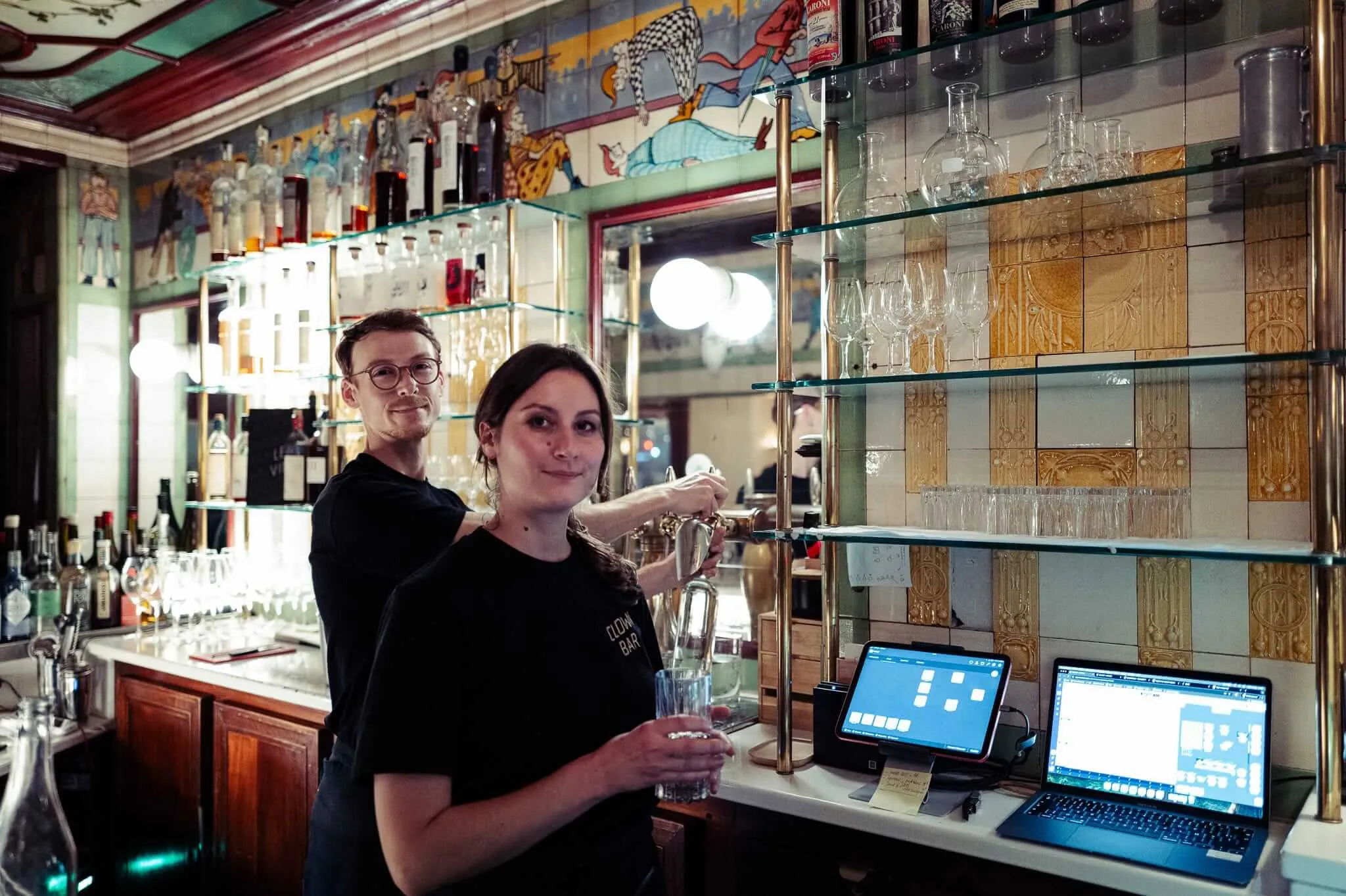 Two bartenders working behind an ornate bar with decorative murals on the walls and ceiling. A woman in black operates a beer tap while a man with glasses stands behind her. Glass shelves display glassware, and laptops are visible on the bar counter, blending traditional pub atmosphere with modern technology.