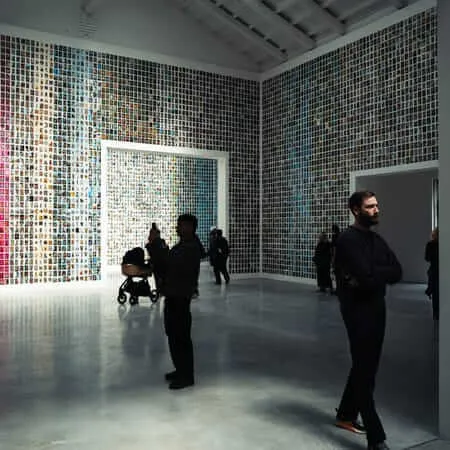 Visitors stand in a large contemporary gallery space whose floor-to-ceiling walls are completely covered in a dense grid of thousands of small images. A bearded man in black stands in the foreground with arms crossed, while a silhouetted figure raises a smartphone to photograph the installation. Further back, visitors with a pram explore the space, and additional rooms visible through doorways reveal more image-covered walls.