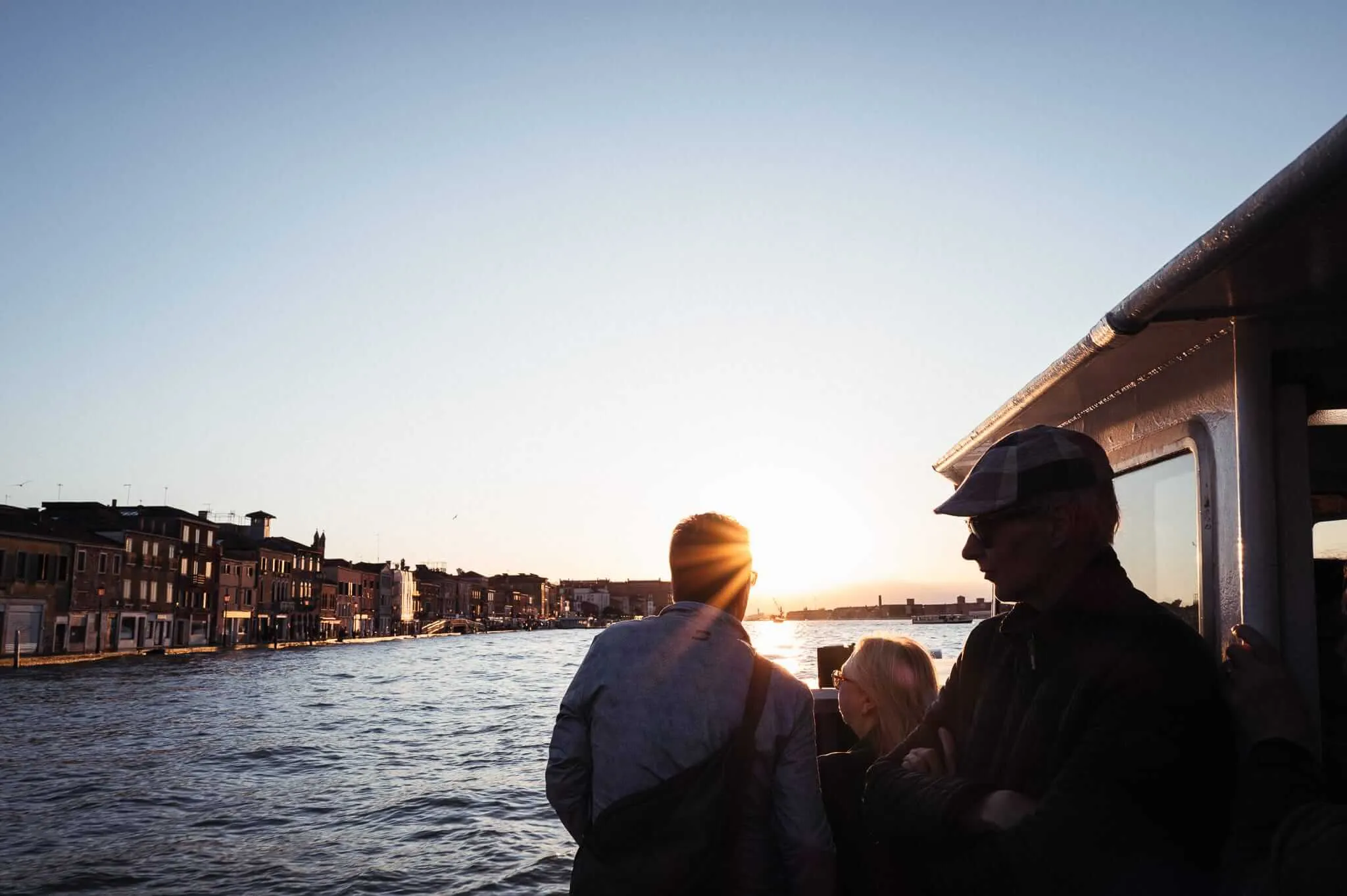 Passengers on a vaporetto water bus silhouetted against a blazing sunset over a Venetian canal, with historic buildings lining the waterway and warm golden light reflecting off the water
