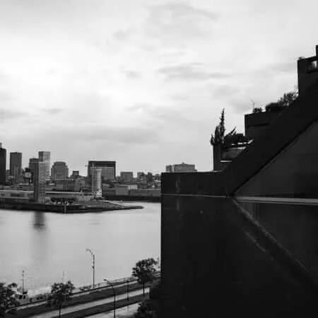 Black and white photograph taken from an elevated position beside a striking brutalist concrete building with angular stepped terraces and rooftop vegetation, looking out across a wide river toward a dense urban skyline under a cloudy sky. A small boat and waterfront promenade are visible in the foreground below.