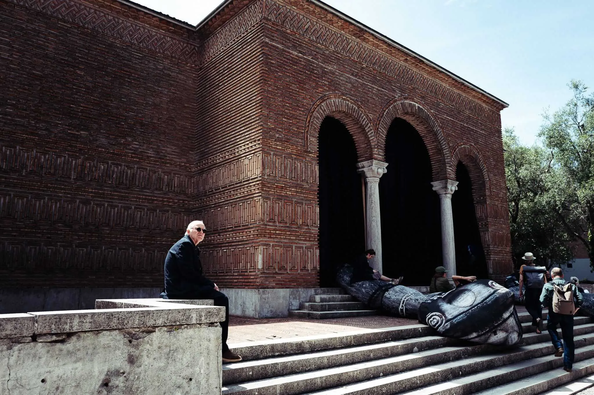 A man in black clothing sits on a stone ledge outside an ornate red brick building with Moorish arched entrances, while a massive dark sculptural figure lies collapsed on the steps nearby, with other visitors examining it in bright afternoon sunlight.