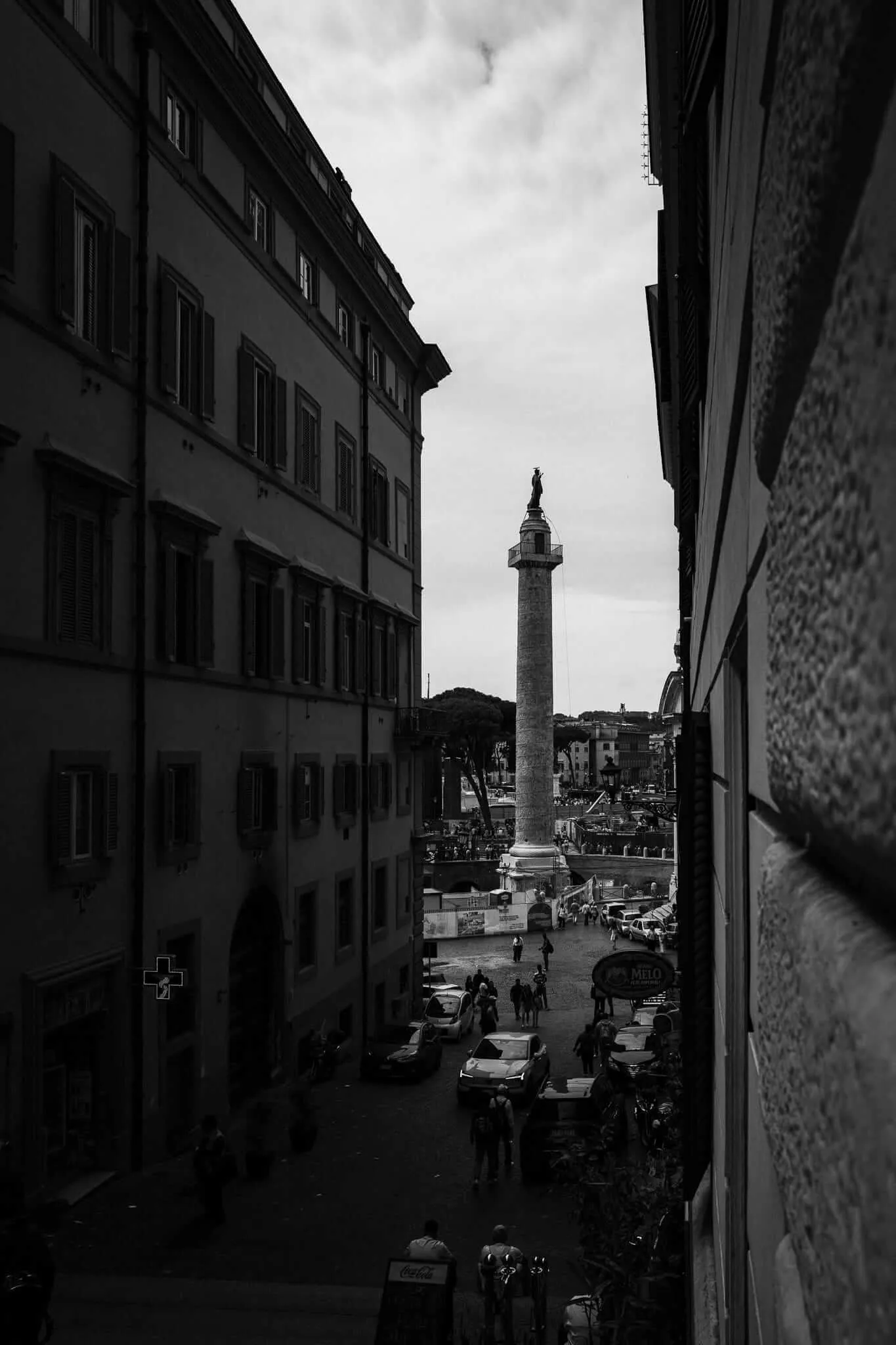 A black and white photograph taken from between two buildings showing a narrow urban view of a tall monument or column with a statue on top, surrounded by historic architecture, parked cars, and people in a European city square