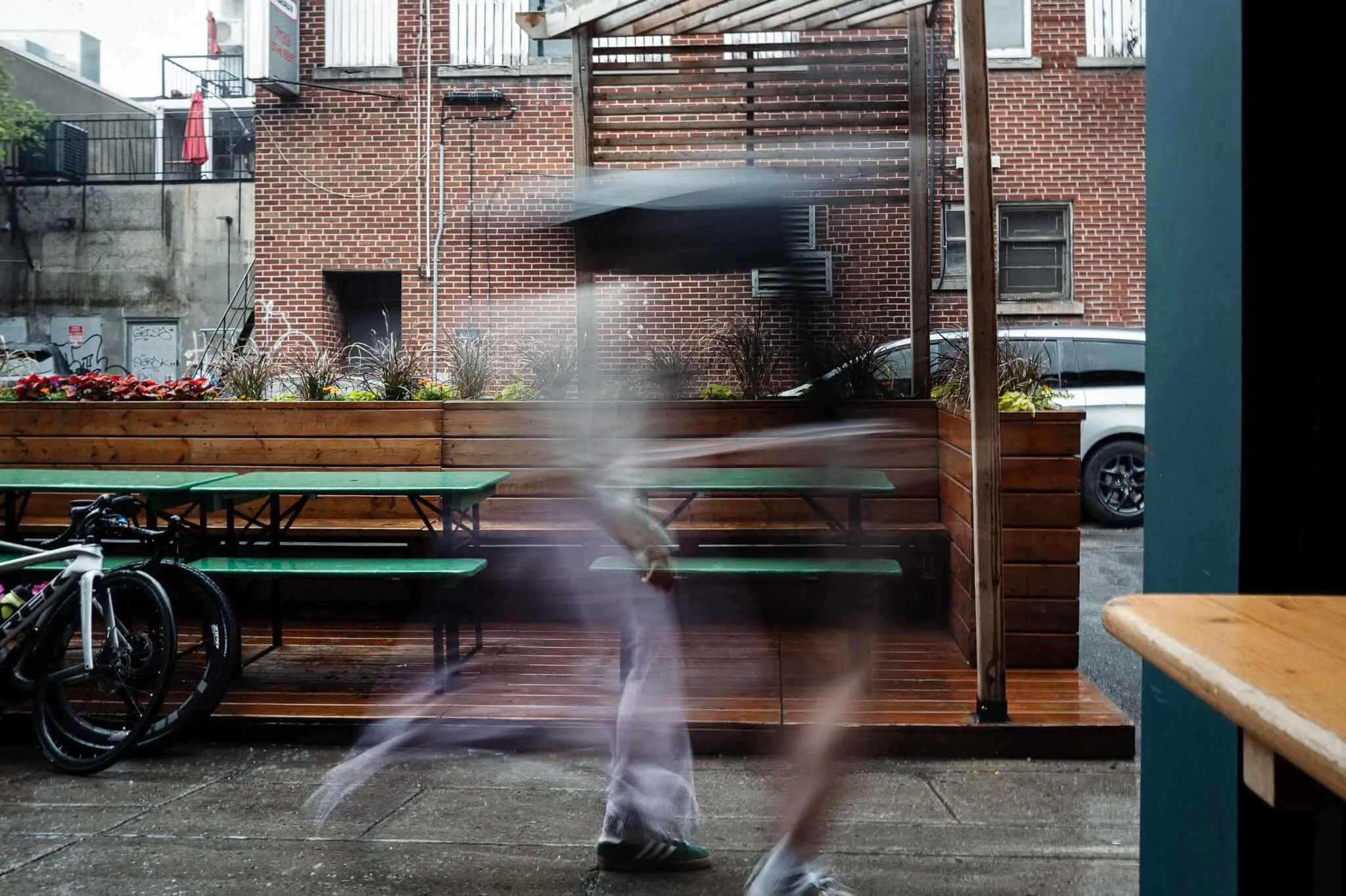 A long-exposure photograph of two people walking past an outdoor restaurant patio, creating a ghostly motion blur effect. Green picnic tables and wooden planters filled with colorful flowers line the deck, with brick buildings visible in the background. A bicycle is parked to the left.