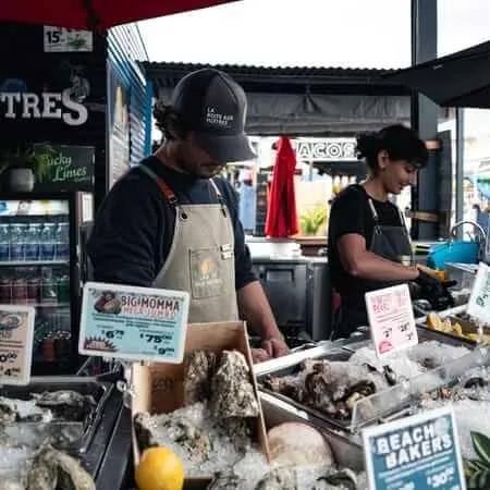 Two workers in aprons shuck oysters at an outdoor market stall called La Boîte aux Huîtres, with trays of fresh oysters on ice and price signs visible in the foreground.