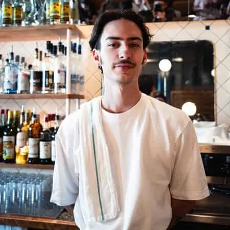 A young male bartender with a mustache and ear piercings stands confidently behind a bar, wearing a white t-shirt with a service towel draped over his shoulder, surrounded by shelves of liquor bottles and warm ambient lighting.