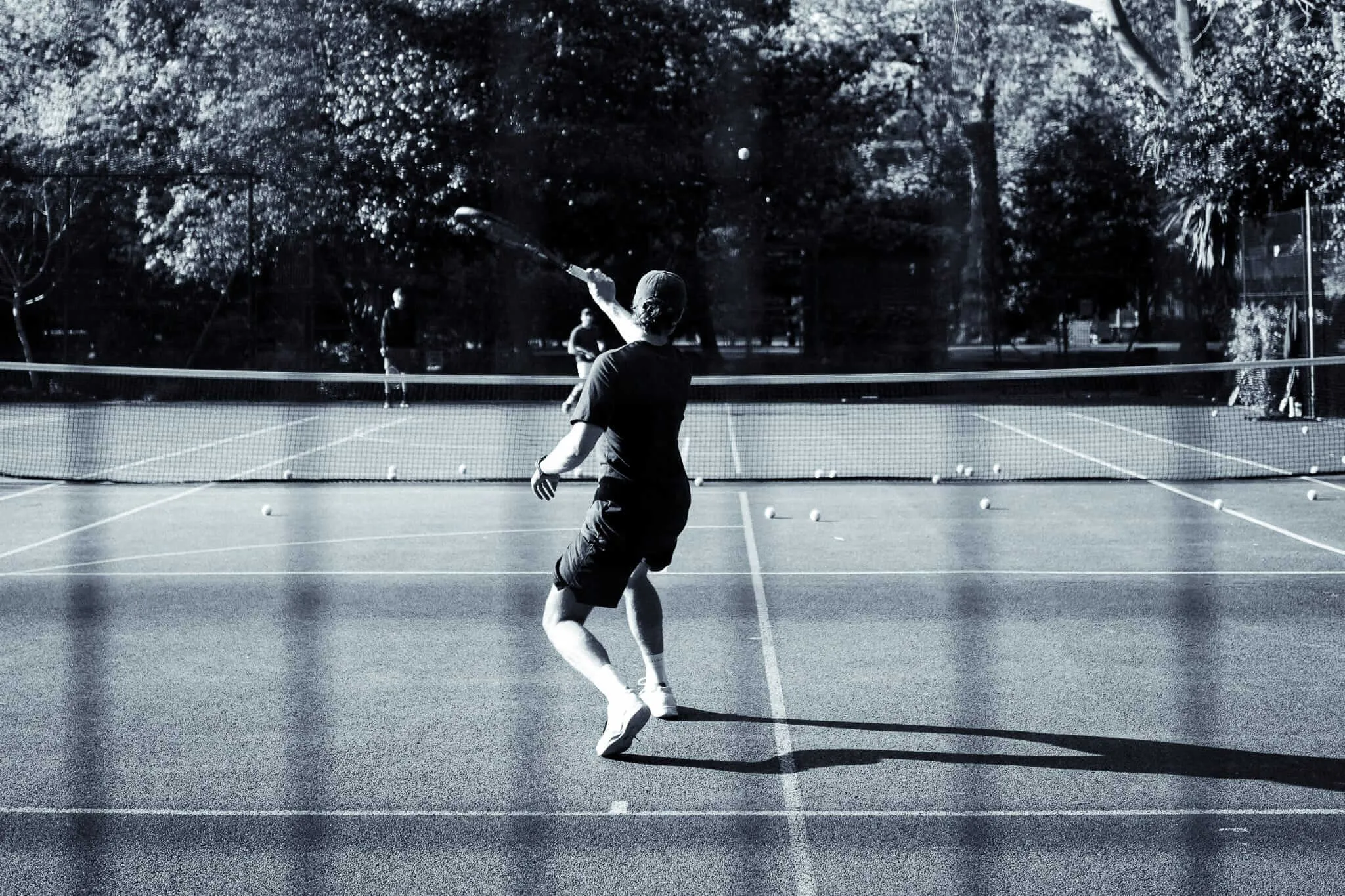 Black and white photograph of a tennis player mid-serve on an outdoor court, captured from behind as they reach up with their racket. The court is surrounded by trees and has tennis balls scattered across the surface, creating dramatic shadows in the natural lighting.