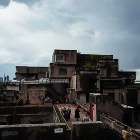 Visitors explore the weathered concrete terraces and walkways of Habitat 67 in Montreal, with a dramatic stormy sky overhead and the St. Lawrence River and port visible in the background.