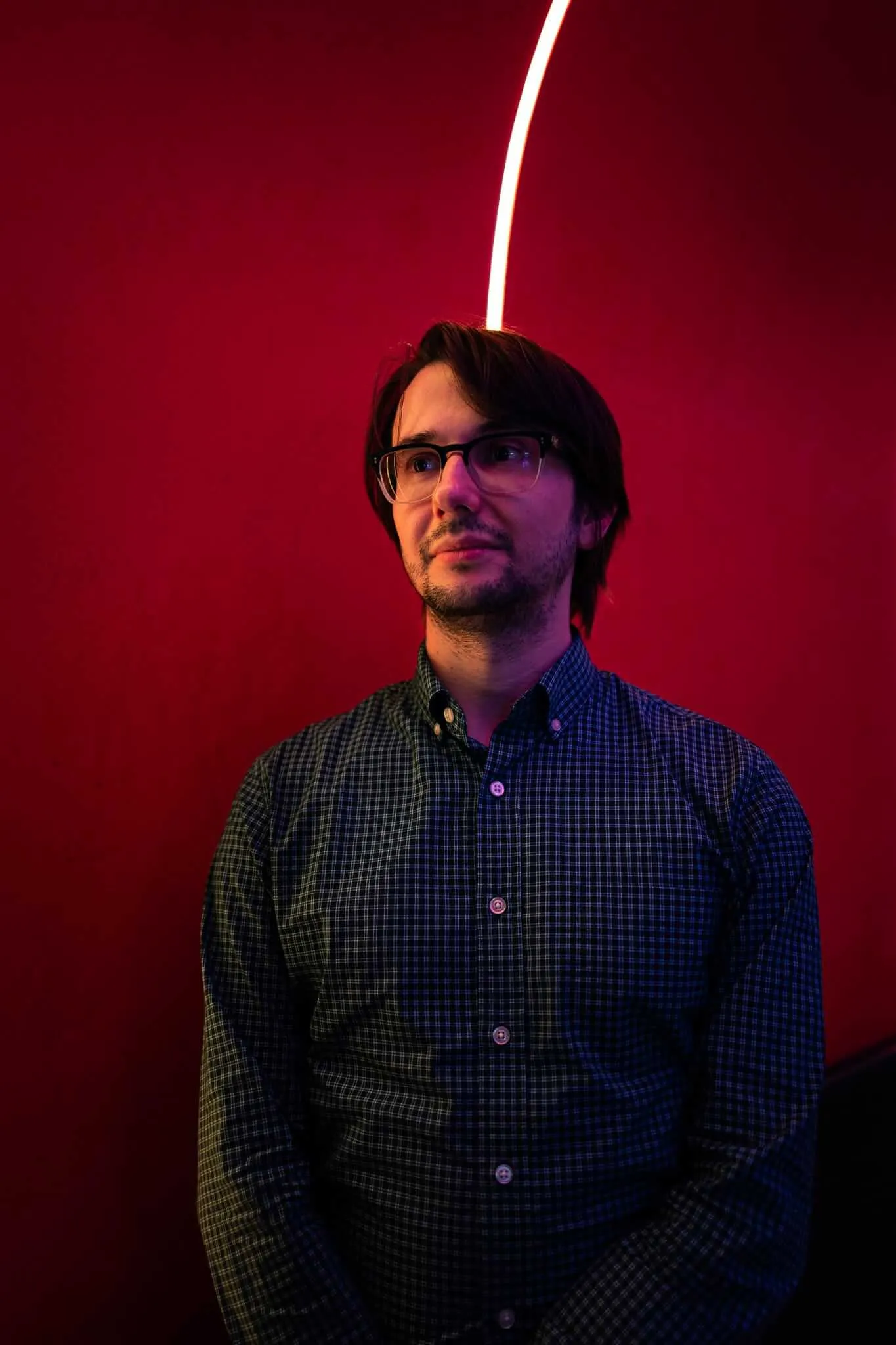 A bearded man with dark-rimmed glasses and dark hair looks upward with a contemplative expression, standing against a deep red background. A curved white neon light arc rises behind his head, creating an ethereal halo effect. He wears a blue and green plaid button-down shirt, bathed in dramatic red and purple light.