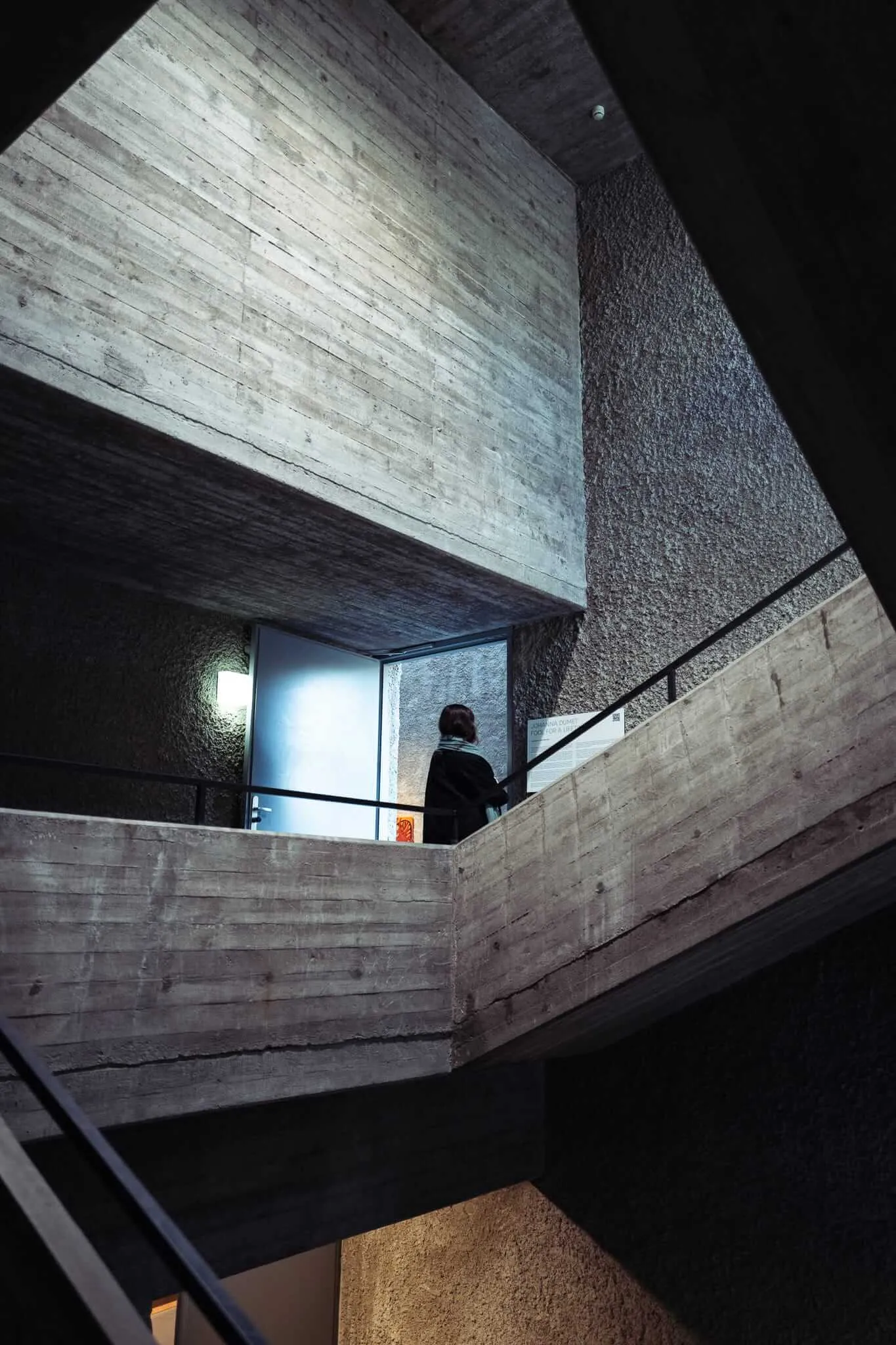 A person in dark clothing ascends concrete stairs in a brutalist architectural space with raw concrete walls, textured surfaces, and dramatic angular lighting from above