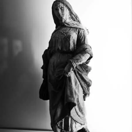 A dark, weathered sculpture of a robed and hooded female figure standing on a circular base, photographed in black and white with dramatic chiaroscuro lighting against a bright white background.