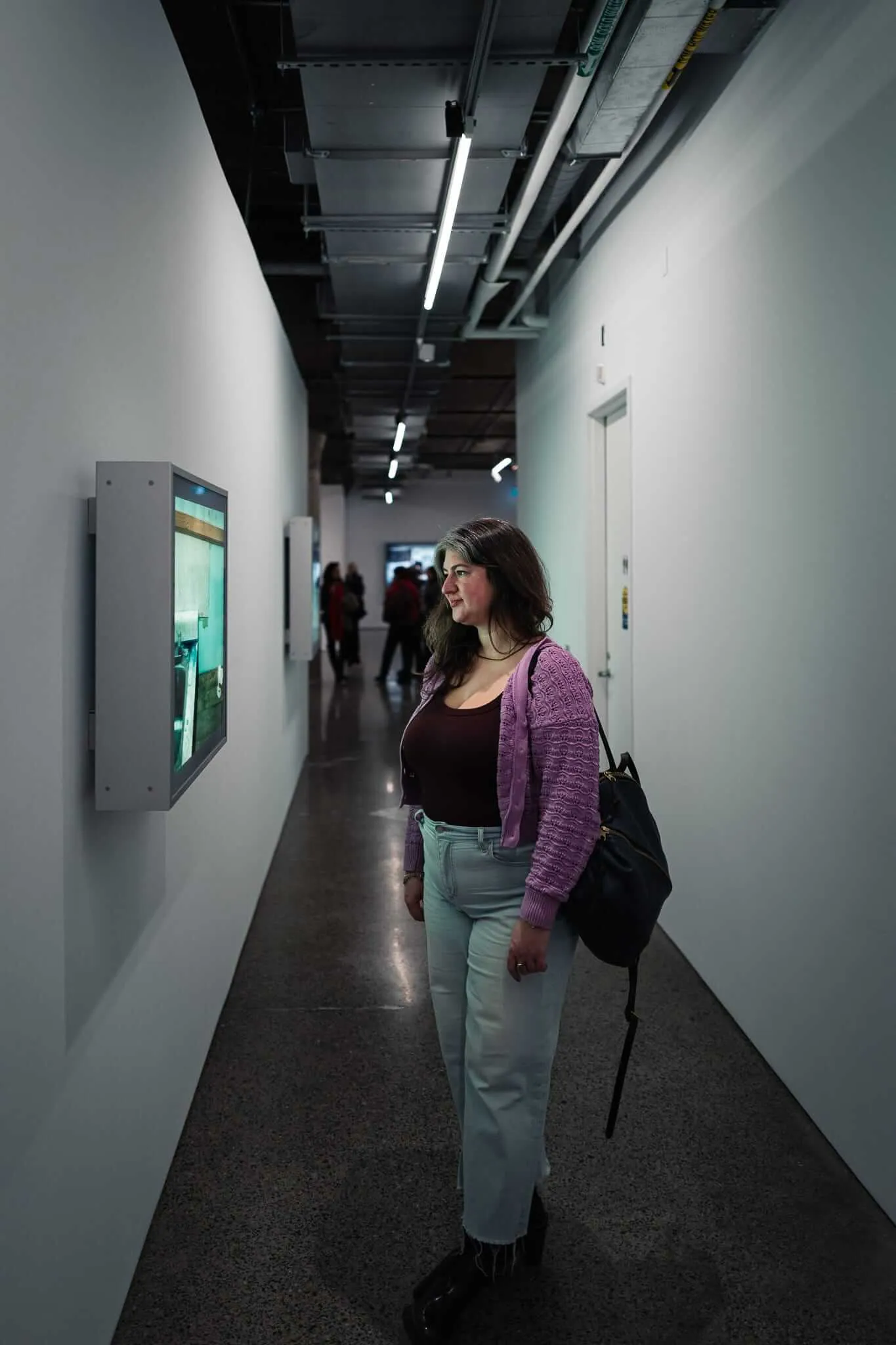 A young woman with dark wavy hair wearing a purple cardigan, dark top, and light jeans stands in a modern institutional hallway with exposed ceiling infrastructure, looking at a wall-mounted digital screen while carrying a dark bag