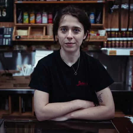 A young woman with dark hair pulled back, wearing a black staff t-shirt and a delicate necklace, leans with arms crossed on a glass deli counter inside a bagel and smoked salmon shop, shelves of jars and bottles visible behind her, looking directly at the camera with a calm, knowing expression.