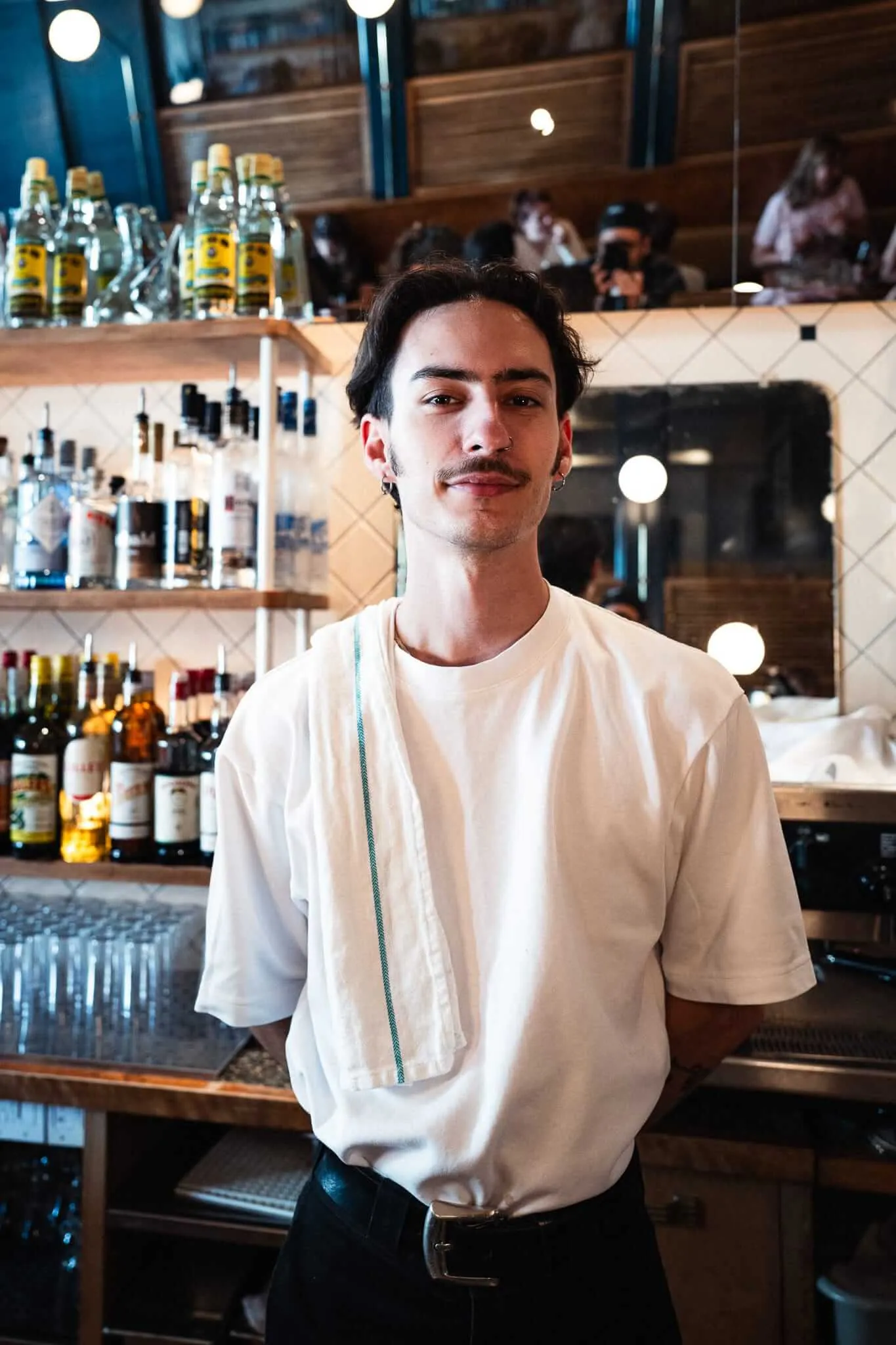 A young male bartender with a mustache and ear piercings stands confidently behind a bar, wearing a white t-shirt with a service towel draped over his shoulder, surrounded by shelves of liquor bottles and warm ambient lighting.