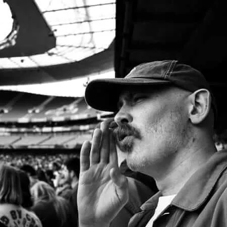 A black and white photograph of a man in a baseball cap cupping his hand around his mouth to amplify his voice while shouting at a football stadium. He has a mustache and goatee, and other spectators are visible in the blurred background. The stadium's distinctive architecture with its curved roof and multiple tiers is visible.