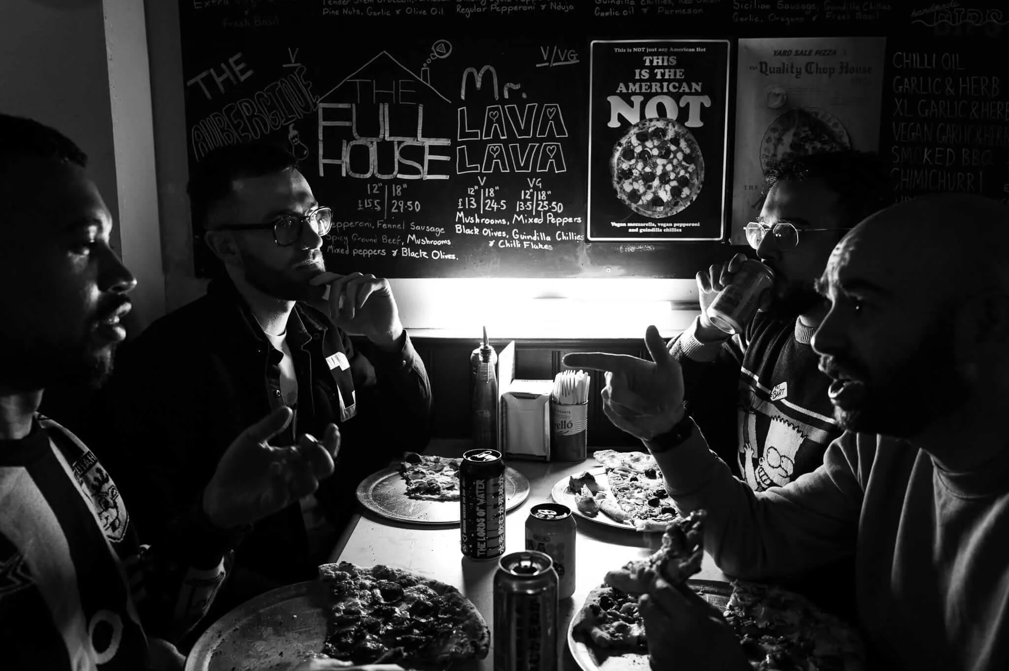 Black and white photograph of four people seated around a table in a pizzeria, with their own pizzas and drinks. Behind them is a chalkboard menu featuring pizza options including 'The Full House' and 'Mr. Lava Lava.' The intimate scene is lit dramatically with strong contrasts between light and shadow.