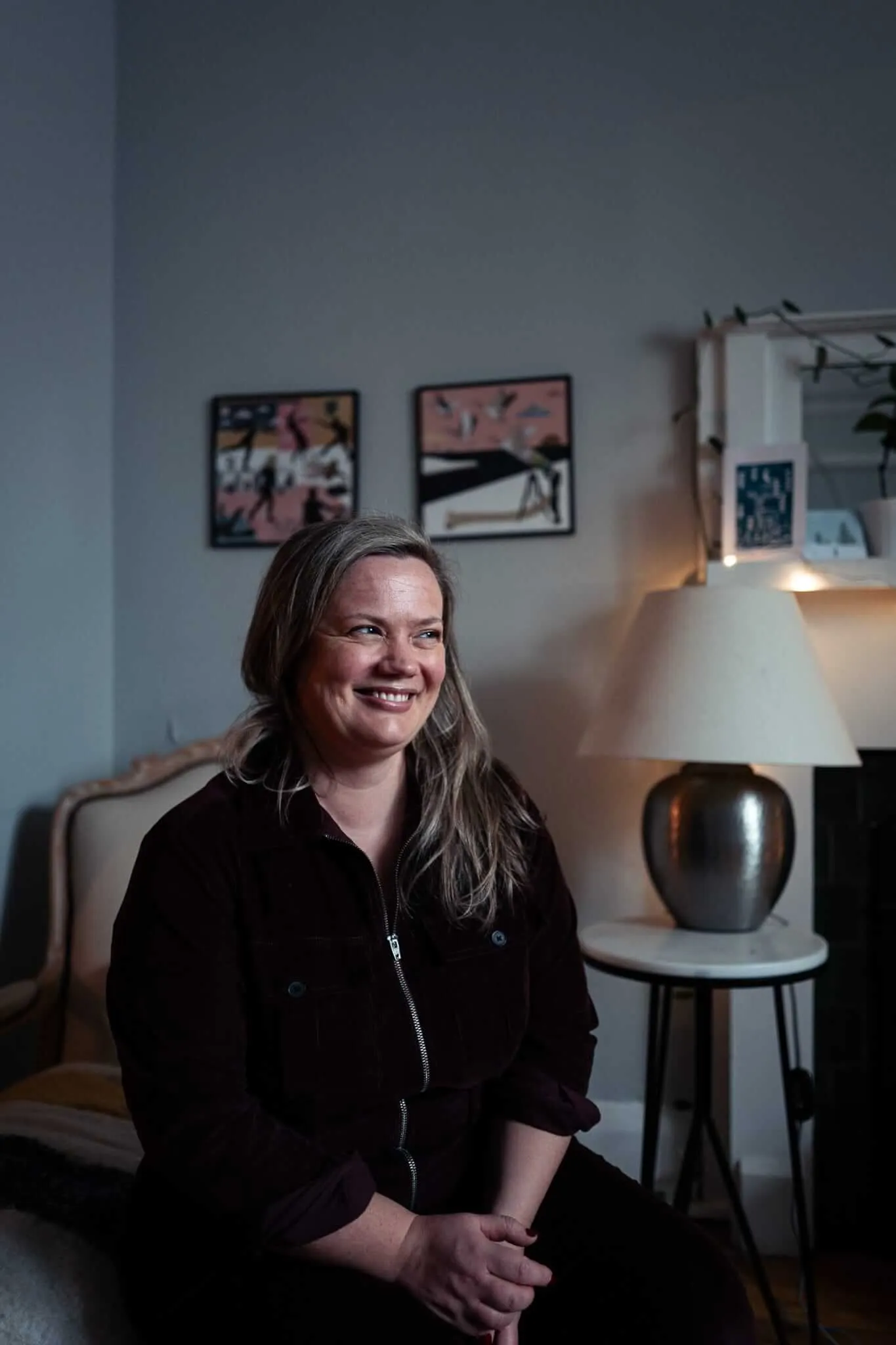A woman with long blonde hair wearing a dark maroon zip-up jumpsuit sits smiling in a modern living room. Behind her are framed artworks on the wall, a cream-colored armchair, and a side table with a distinctive lamp featuring a white shade and dark metallic base. The room has gray walls and warm lighting.