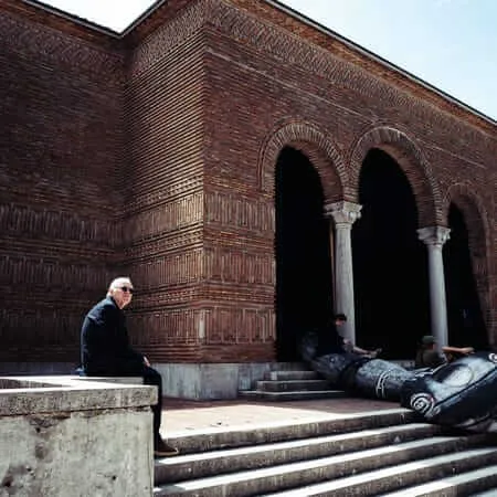 A man in black clothing sits on a stone ledge outside an ornate red brick building with Moorish arched entrances, while a massive dark sculptural figure lies collapsed on the steps nearby, with other visitors examining it in bright afternoon sunlight.