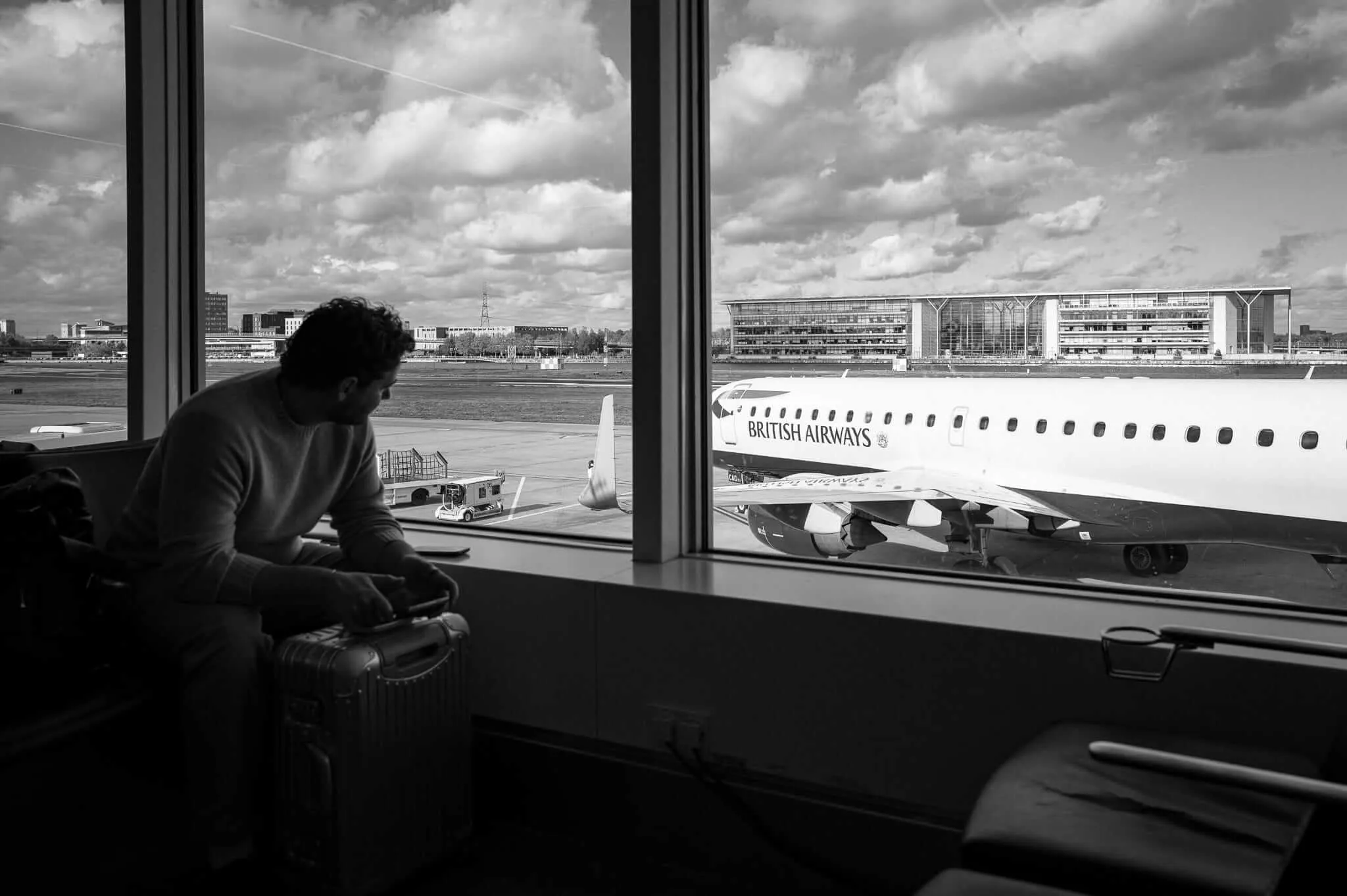 Black and white photograph of a person sitting alone in an airport terminal, looking out through large windows at a British Airways aircraft on the tarmac. The person appears contemplative, positioned in silhouette against the bright exterior view of the plane, runway, and airport buildings under a cloudy sky.