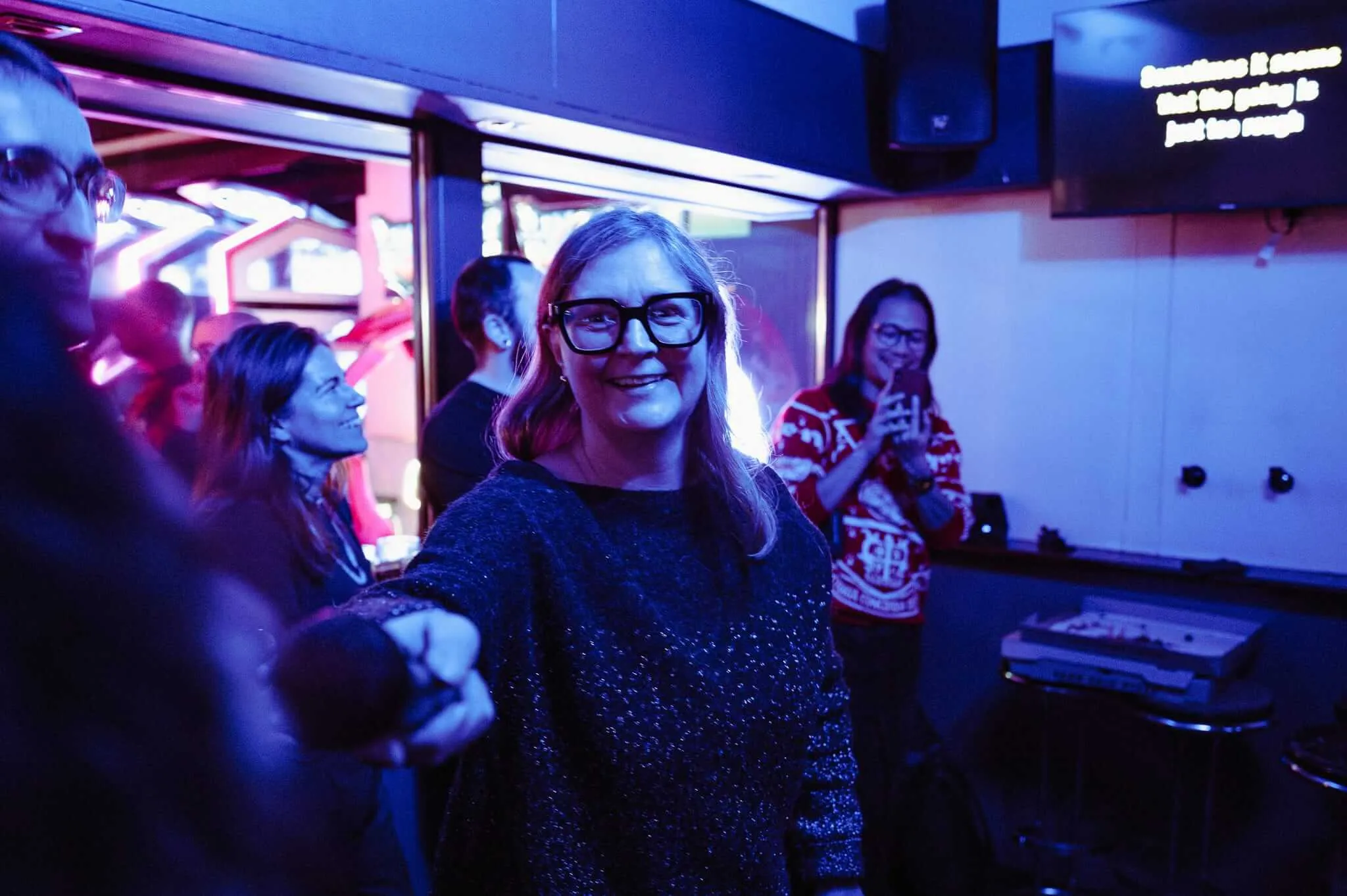 A woman with glasses and long hair wearing a sparkly dark sweater smiles warmly in the foreground of what appears to be a karaoke room or bar, bathed in purple and blue lighting. Behind her, other people are visible including someone in a patterned sweater, with illuminated display screens and entertainment equipment visible in the background.