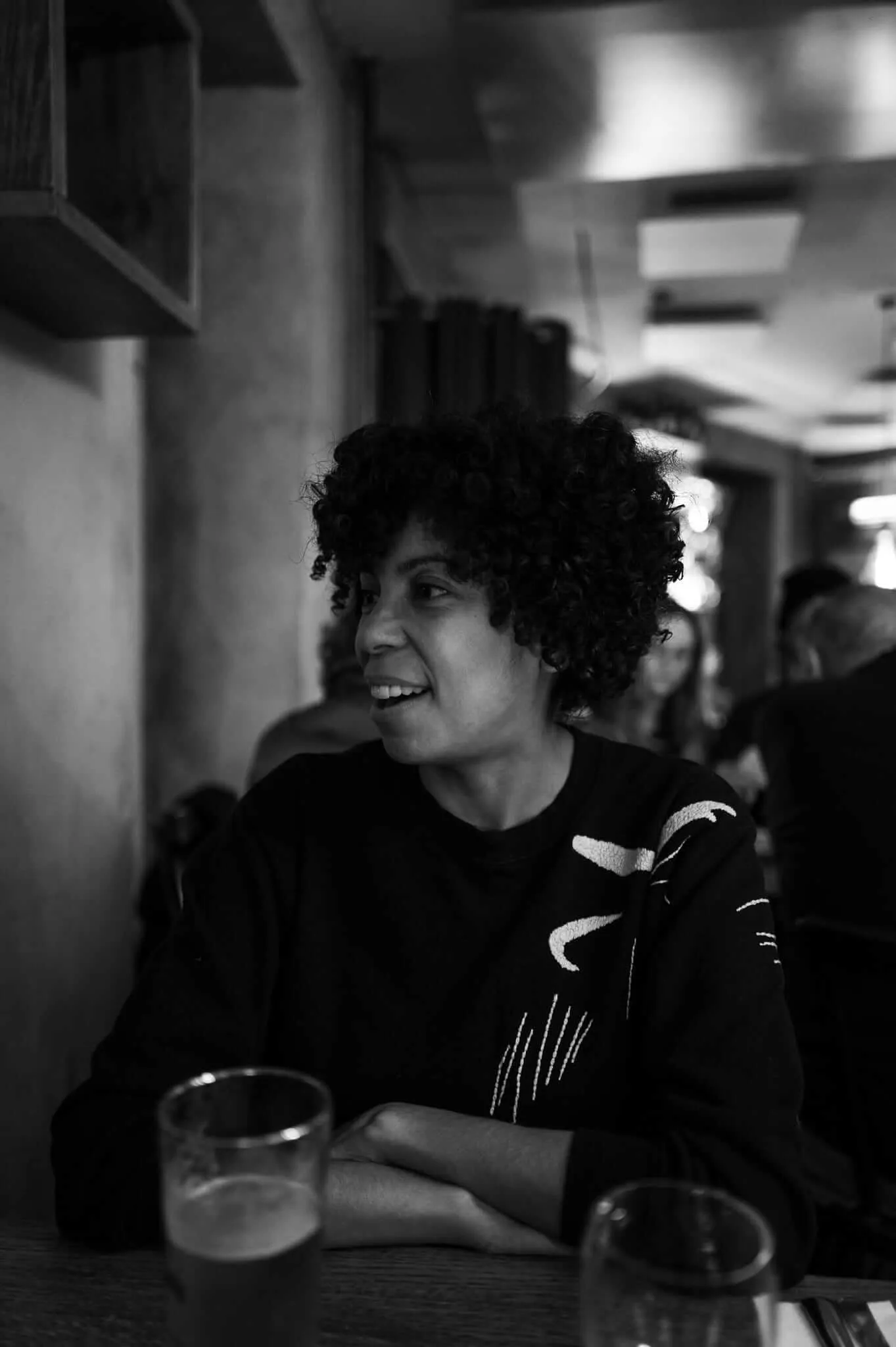 Black and white portrait of a person with curly hair sitting at a table with glasses, smiling warmly while looking to the side in what appears to be a restaurant or cafe setting