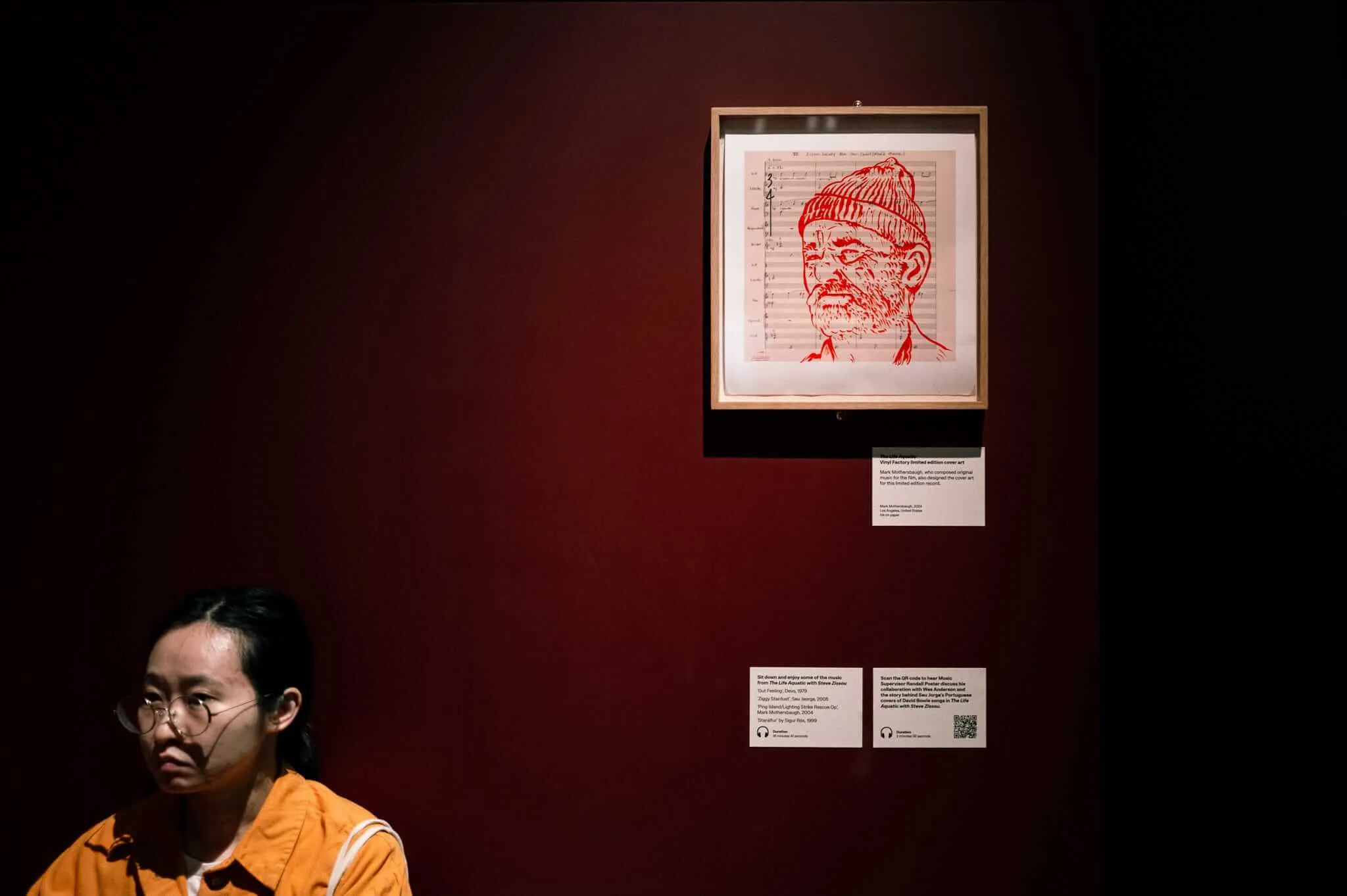 A person wearing glasses and an orange shirt sits in contemplation in front of a dark red museum wall, beneath a framed red sketch portrait of a bearded man in a knit cap (Bill Murray as Steve Zissou) drawn on sheet music
