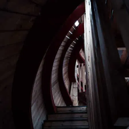 A dramatic low-light view looking up through a wooden spiral staircase with dark red curved railings, receding into a dim tunnel-like structure with minimal lighting visible at the top