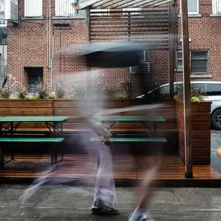 A long-exposure photograph of two people walking past an outdoor restaurant patio, creating a ghostly motion blur effect. Green picnic tables and wooden planters filled with colorful flowers line the deck, with brick buildings visible in the background. A bicycle is parked to the left.