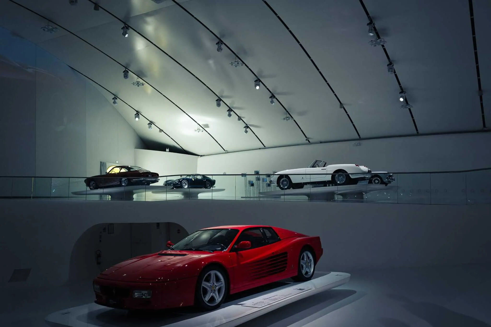 A red Ferrari sports car displayed on a white platform in the foreground of a sleek museum gallery, with several classic cars arranged on an elevated mezzanine level behind a glass partition, all illuminated by dramatic track lighting against curved white walls.