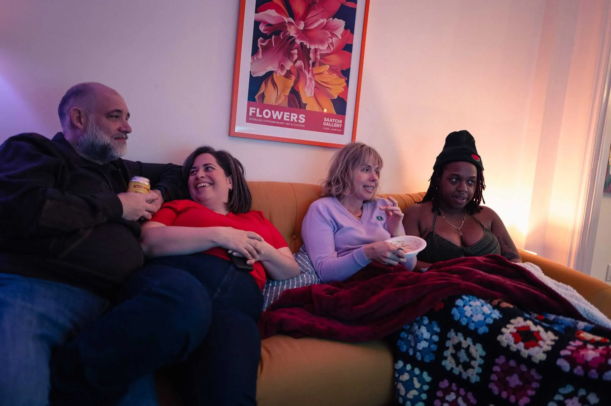 Four people sitting together on a tan leather couch in warm ambient lighting. From left: an older bearded man in dark clothing, a woman in red, a blonde woman in light blue holding a small bowl, and a Black woman in dark clothing with a beanie. They're covered by blankets including a colorful crocheted throw. A floral art poster hangs on the wall behind them.