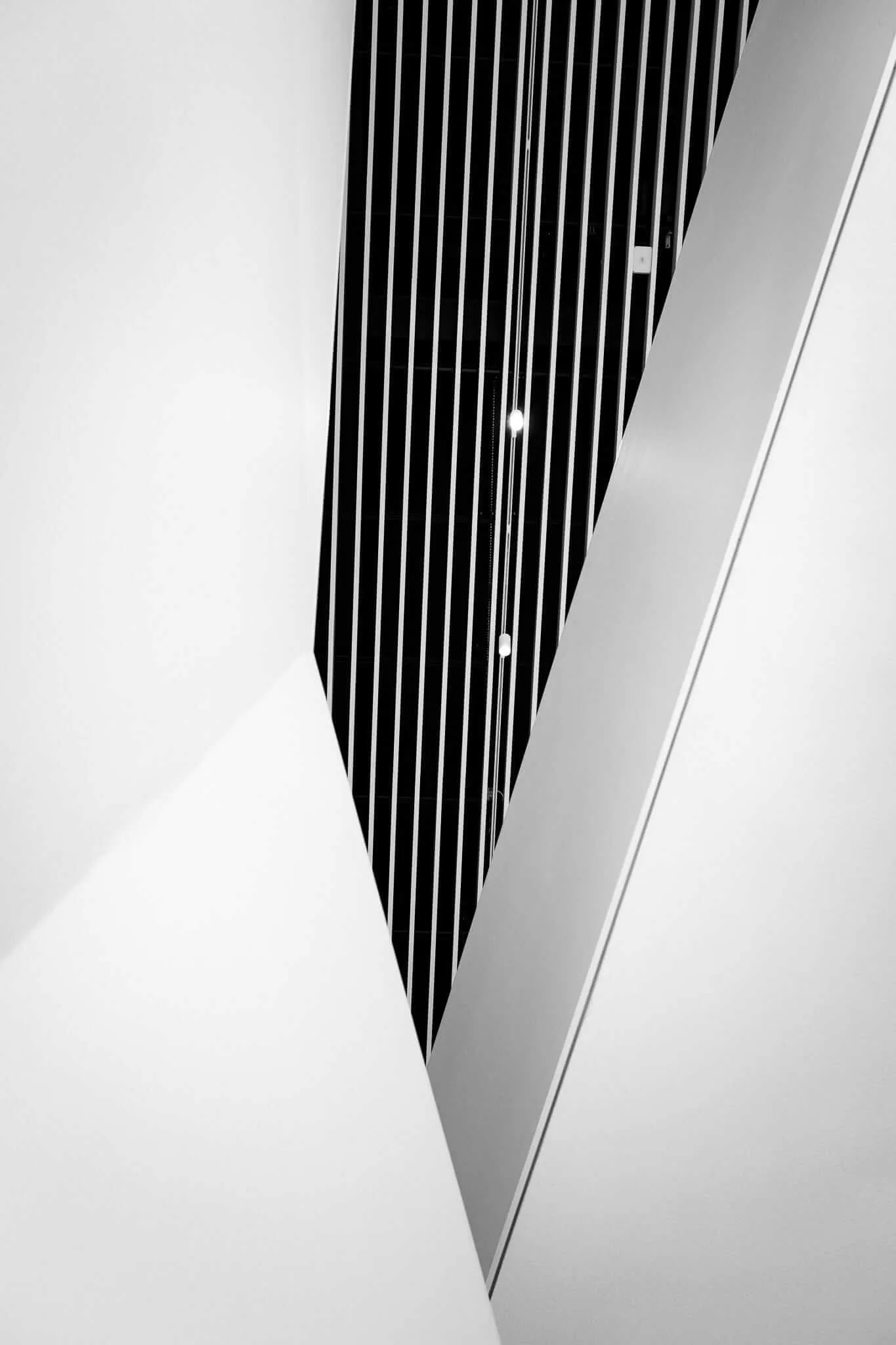 Black and white architectural photograph showing a dramatic upward perspective of a building with vertical metal slats or railings forming strong parallel lines against white walls, creating a sharp V-shaped composition with geometric shadows