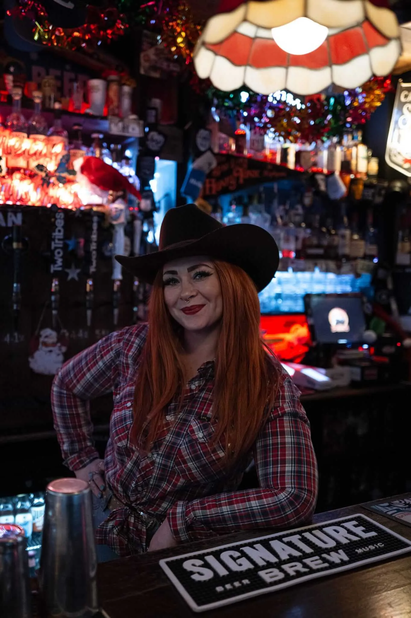 A woman with long red hair wearing a black cowboy hat and red plaid shirt sits at a bar counter in a dimly lit honky-tonk establishment. The background features colorful neon lights, a Tiffany-style lamp, and various bar memorabilia creating an authentic dive bar atmosphere.