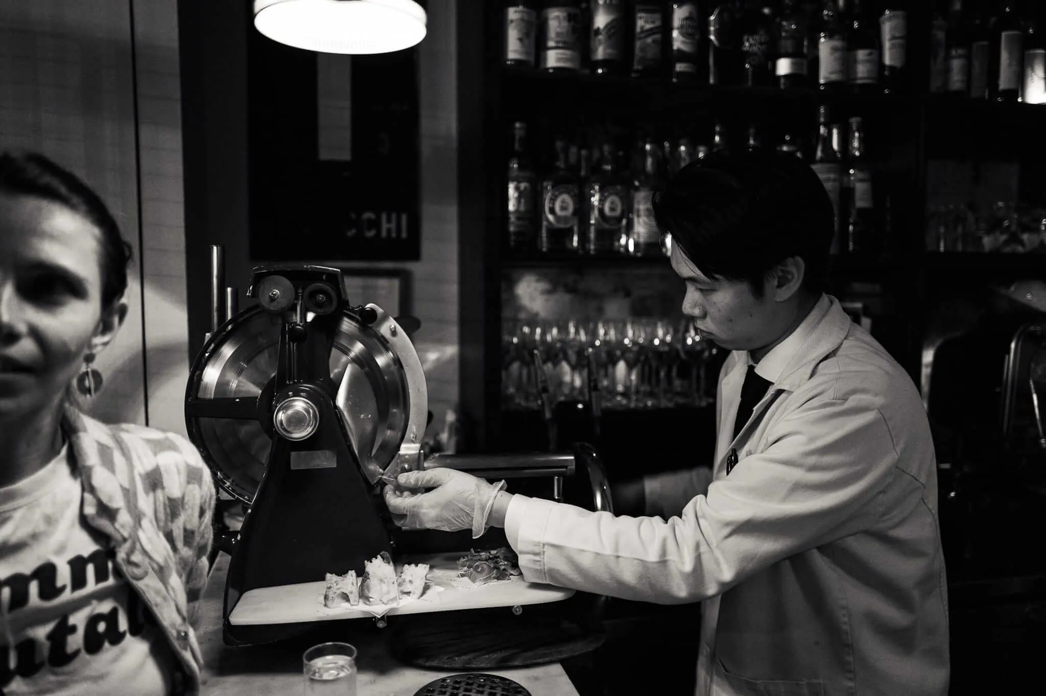 Black and white photograph showing a person in white chef's coat operating a vintage meat slicer in what appears to be a restaurant or delicatessen, with bottles lining shelves in the background and another person visible on the left side of the frame