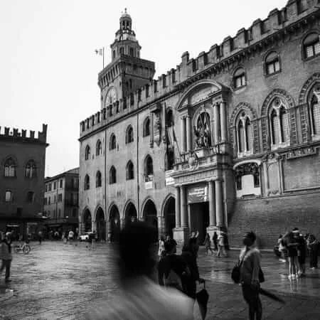 Black and white photograph of Bologna's Palazzo d'Accursio and Palazzo del Podestà in rainy weather, with a ghostly motion-blurred figure in the foreground and scattered pedestrians crossing the wet cobblestone piazza beneath a grey sky
